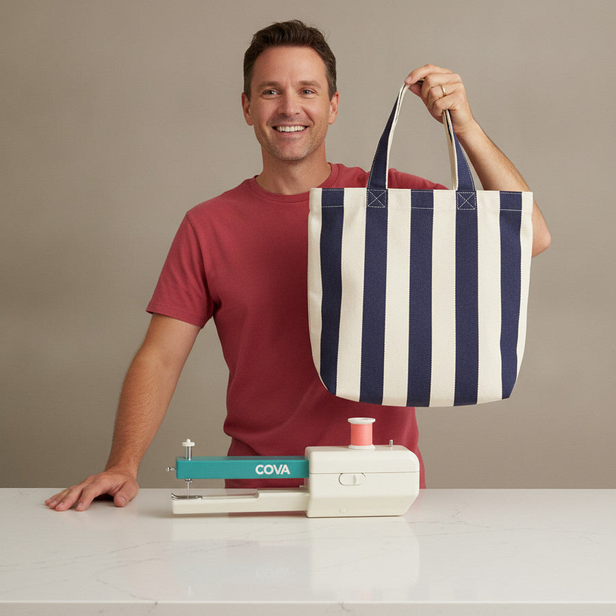 A man in a red t-shirt holding a striped tote bag he sewed, the COVA handheld machine on the counter